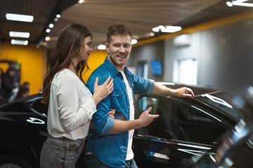 young couple handsome man and beautiful woman looking for new vehicle in electric car dealership center