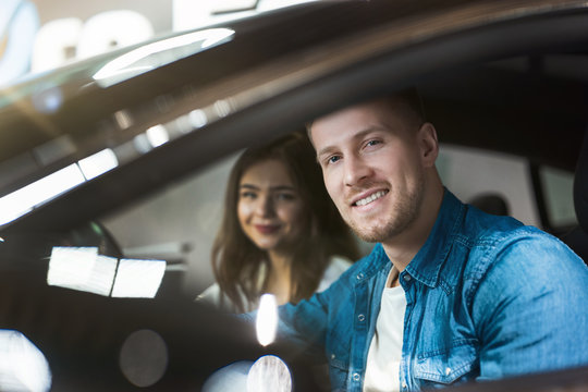 young couple handsome man and beautiful woman checking new vehicle interior in electric car dealership center, looking excited, happy moments