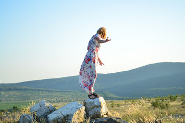 On a hill of stones, a girl stands balancing and looks into the hilly distance covered with a haze.
