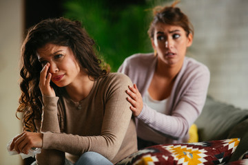 Woman broke up with boyfriend, she is crying and sister is trying to calm her down. Beautiful women sitting in living room. 