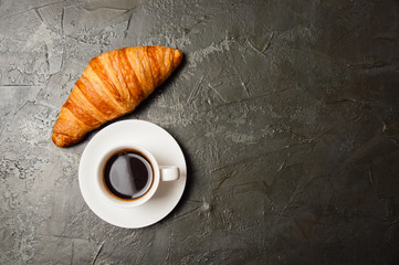 Coffee in white cup and croissant on a dark concrete background, top view, flat lay. Concept for breakfast, coffee break or business lunch. There is a place for text on the right