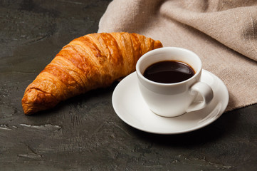 Croissant and cup of coffee and saucer on dark background with a gray linen napkin