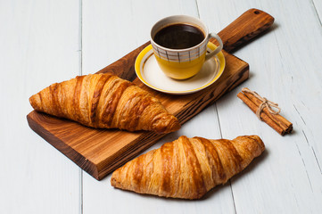 Two tasty croissants on wooden board and coffee in a vintage yellow cup and saucer on a light background