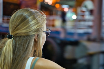 blonde girl with back glasses facing the front at night with the lights of the buildings around