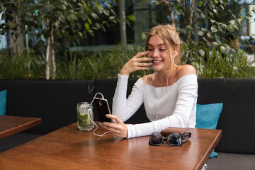 Cheerful blond woman blogger with cocktail is reading incoming sms message on smartphone connected to free wifi in cafe. Positive young student watching broadcasting online on modern mobile phone.