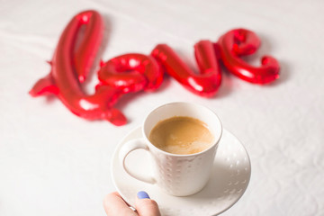 A cup of coffee in a female hand on the background of a red foil balloon in the shape of the word love. Valentine's day concept.