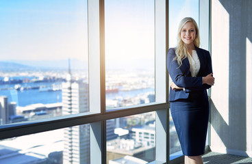 Smiling young businesswoman standing by office windows overlooking the city