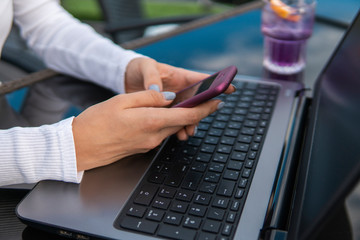 Modern business woman is reading incoming sms message on smartphone or watching broadcasting online on modern mobile phone while sitting in caffe with a laptop. Female student sitting in coffee.