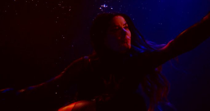 A beautiful femme fatale with unusual makeup and long hair is in a dark Studio under water on a blue flashing background. Her hair is flying.