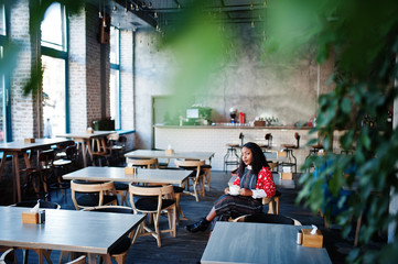 Сonfident young african american woman in smart casual wear at cafe with cup of hot drink in hand.