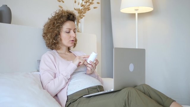 Pregnant Woman Using Laptop And Reading Label On Medicine Bottle At Home