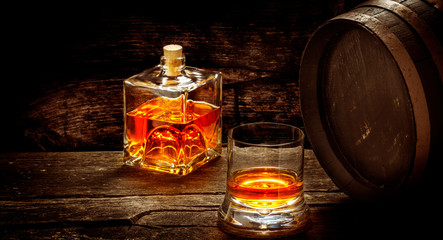 Whiskey bottle and whiskey glass stand on a wooden table in front of a whiskey barrel, still life in the distillery