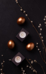 copper colored Easter eggs on a black background next to willow branches and candles. dark style