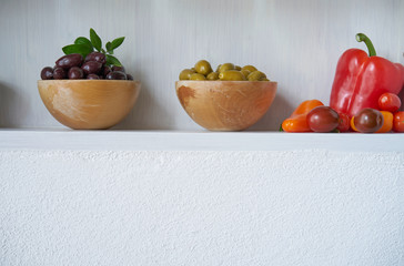 still life olives green brown and black in bowl with vegan red vegetables like tomatoes and paprika, white wall copy space vitamins