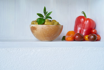 still life olives green in bowl with vegan red vegetables like tomatoes and paprika, white wall copy space vitamins