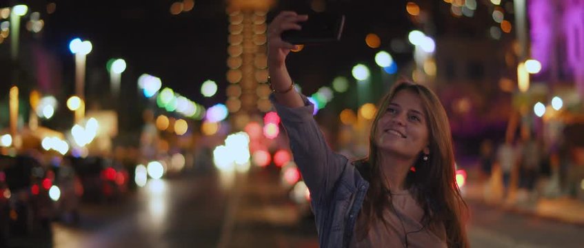 A young woman taking a selfie in the middle of the street, night city bokeh lights. Shallow dof, realtime, BMPCC 4K