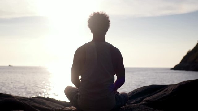 Back view of man sitting in lotus position and meditating at sunset by the sea