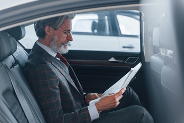 Modern stylish senior man with grey hair and mustache reading newspaper inside of the car