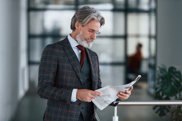 Senior businessman in suit and tie with gray hair and beard holds newspaper in hands