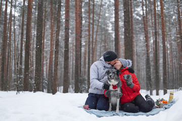 Fototapeta na wymiar young couple with dog have picnic in snowy forest, man and woman kiss