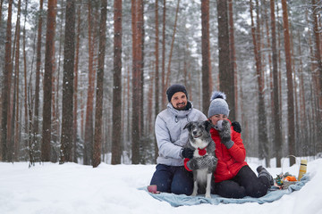 young couple with dog have picnic in snowy forest