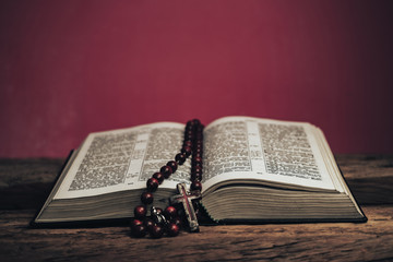 Open Holy Bible and beads crucifix on a old oak wooden table. Beautiful red wall background.