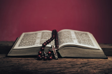 Open Holy Bible and beads crucifix on a old oak wooden table. Beautiful red wall background.