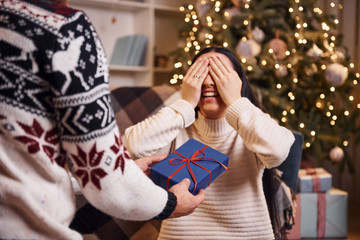 Man covers eyes of hir girlfriend and giving new year surprise in christmas decorated room