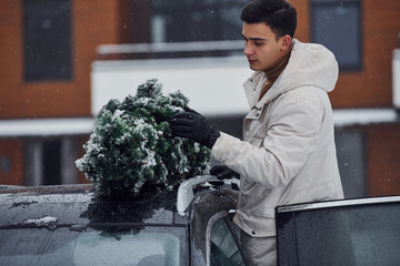 Man in white wear putting christmas fir tree on the top of his car outdoors