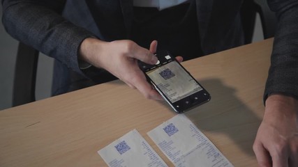 American businessman entering code ising smartphone sitting at table in office room. Young man employee holding phone in hand and scan qr, working at desk in company. Entrepreneur wearing classic - Powered by Adobe
