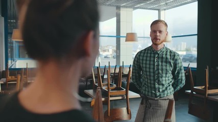 Portrait of young bearded waiter standing in empty restaurant and talking to owner