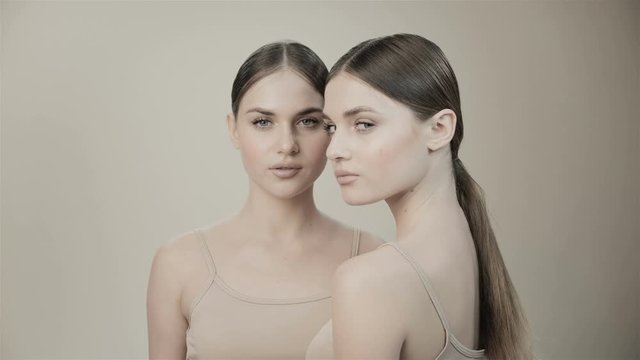Two twin girls in with nude makeup are looking at the camera. In the studio on a beige background. Natural beauty. Sisters. Skin care cosmetics.