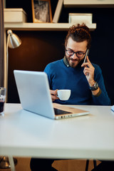 Young smiling caucasian bearded blogger sitting in his office, talking on the phone, holding cup of coffee and taking a break from work.