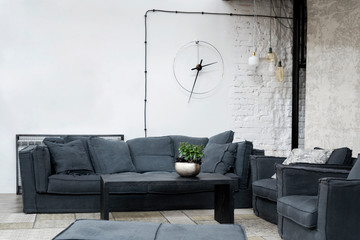 Industrial interior of living room in loft apartment with white brick wall and grey modern sofa. Spacious room with stylish furniture. Clock on the wall.