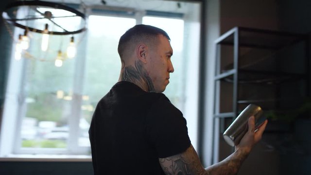 Tracking arc shot of young tattooed waiter showing tricks with cocktail shaker and flipping it, 360 degree