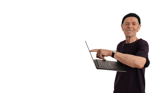 Happy Old Man Pencioner In Black Hat And T-shirt Pointing At Blank Screen Of Laptop Computer And Looking At The Camera, Holds Laptop Computer In His Hand And Smiling Isolated Over White Background