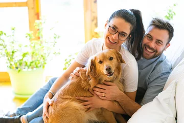 Fotobehang Happy couple with a golden retriever dog sitting on a sofa smiling and positive © gabriele