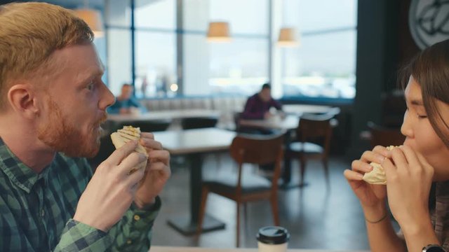 Man eating doner kebab with his asian girlfriend sitting in typical city junk food bistro cafe. Midlle eastern fast food cuisine at lunch time