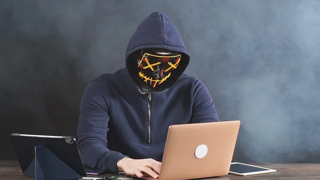Young programmer in smoke-filled background, hacker in mask, trying to hack into a computer system, unrecognizable male sit using laptop