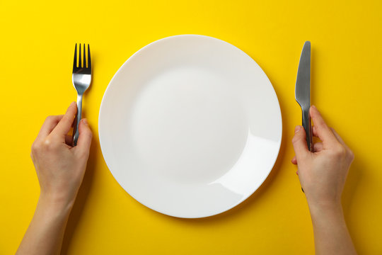 Female hands hold fork and knife on yellow background with plate, top view