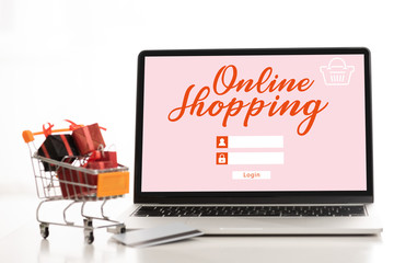 Laptop with online shopping lettering, credit card with toy gifts in cart on table on white background