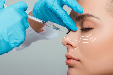 cropped view of cosmetologist making beauty injection in nose to attractive woman isolated on grey