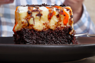 A piece of chocolate cake with custard, whipped cream, chocolate flakes and a drop of orange glaze lies on a plate