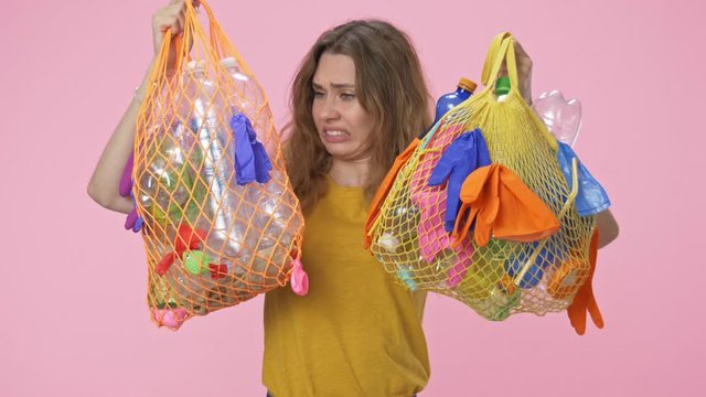 Displeased young woman is holding string bags with plastic waste screaming to the camera isolated over pink background