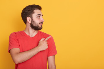 Horizontal shot of smiling young handnsome Caucasian man pointing to empty space aside with his index finger, wearing casual clothing, over yellow wall. Copy space for advertisment or promotion