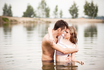 Fototapeta premium Young beautiful loving couple standing in still water and looking at each other on summer day with green nature at background. Love, relationships, dating, flirt and attraction concept