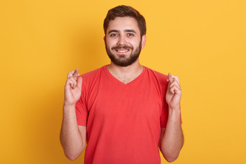 Indoor shot of glad positive bearded man crosses his fingers, anticipate hearing good news, handsome guy posing isolated over yellow background, happy young male rejoices his dreams came true.