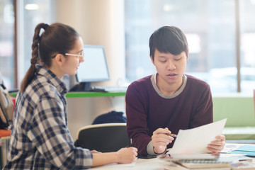 Obraz premium Asian young man reading a report and discussing it together with young woman while they sitting at the table in the classroom