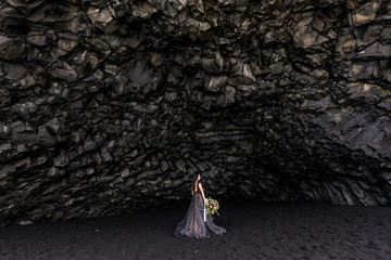 bride in black clothes stands in a black cave on a black sand beach near the Atlantic Ocean in...