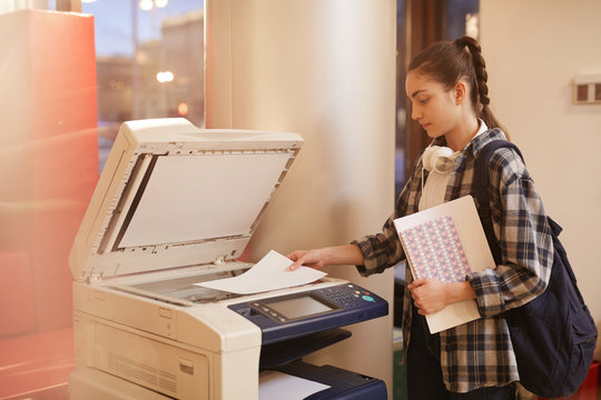 University student with backpack behind her back holding report and making copy of it on the printer